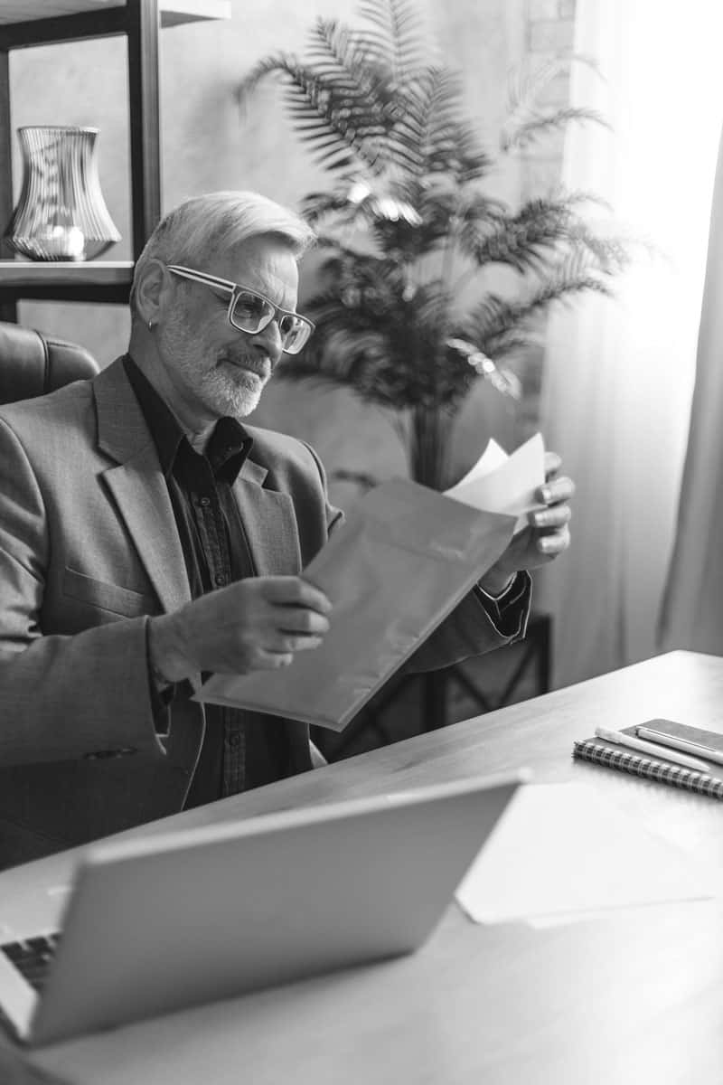 A businessman sitting on a chair with documents in his hands at the table and a laptop on top